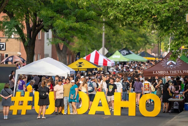 A yellow sign that says #UIDAHO is displayed. The sign is surrounded by tents and a large group of students.