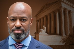 A black man with a beard wearing a suit stands in front of the Supreme Court building