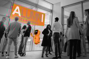 A gathering of people under a banner that highlights the "a" and "i" in the word "alumni" with a robot among the crowd.