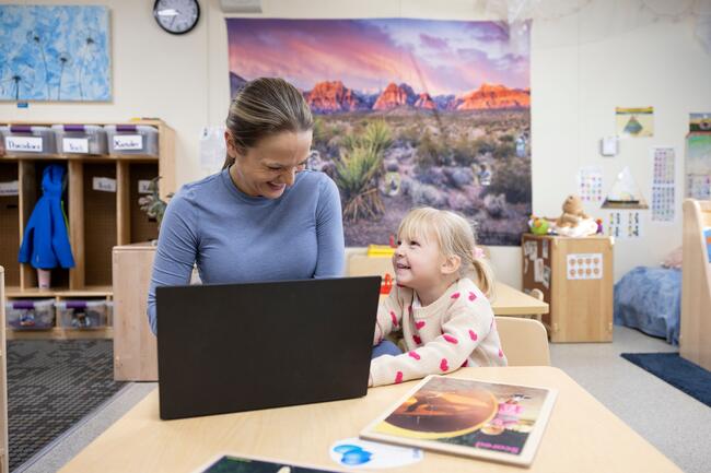 Mother working on her laptop during drop-off as her daughter tries to get her attention at a childcare center on a college campus.
