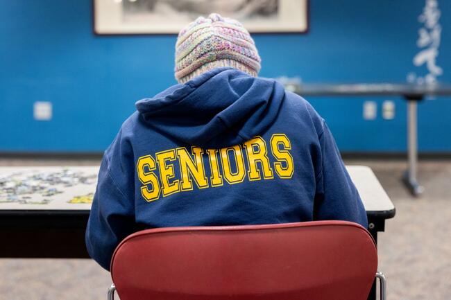 Student working on a puzzle in a common area on a college campus with the word "SENIORS" across their back.