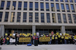The large Department of Education building can be seen behind rows of protesters holding signs reading, "Fund schools not billionaires" and "Trump: Stop stealing from kids."