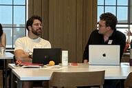 Two men work at laptops in conversation