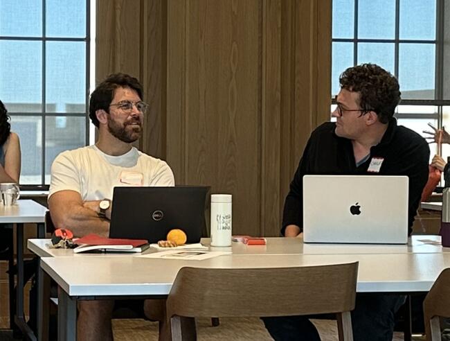 Two men work at laptops in conversation
