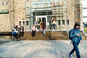 Medium group of multiethnic students going in and out of a college building entrance. Some are sitting and standing on a small staircase on a sunny day.
