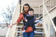 Millennial mother university student walking down housing stairs holding books with infant in a front carrier.