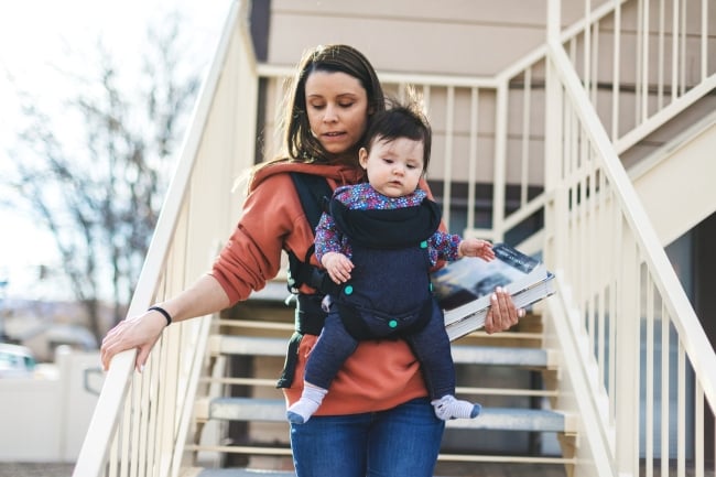 Millennial mother university student walking down housing stairs holding books with infant in a front carrier.