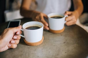 A close-up image of two people holding mugs of coffee on a table, as if at a coffee shop.