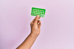 Young brown hand holding birth control pills over isolated pink background.