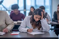 A young woman sits at her desk and takes notes. She has her head in her hand in concentration.
