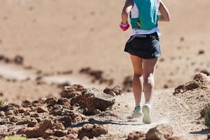 A photo from behind of a woman running on a mountain trail, wearing a hydration vest.