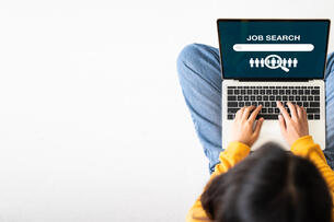 A young adult woman dressed casually in jeans and a sweater sits cross-legged on the floor, with a laptop open to a page that says "Job Search." The photo is taken from overhead.