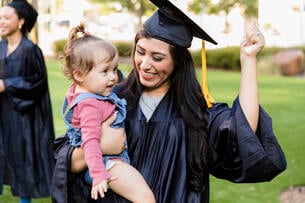 A woman in a graduation cap and gown holds a child and smiles.
