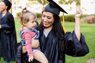 A woman in a graduation cap and gown holds a child and smiles.