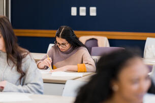 A female student sits in the back row of a classroom, working diligently on an assignment; she is using an eraser to correct an answer.