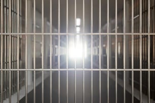 A picture of a dark prison corridor, with a set of bars in the forefront of the picture, with light shining through in the back.
