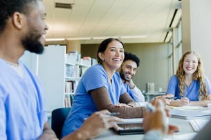 A group of four students in light blue scrubs sit at a table, smiling. 