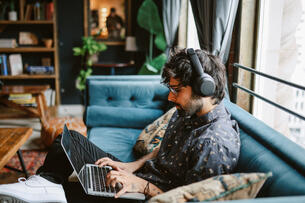 A photo of a young professional working from a couch at home, wearing headphones and working intently at a laptop.