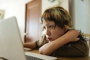 A young Asian American woman searches for jobs on her laptop; she is in deep concentration as she leans down to stare at the computer while resting her head on one of her arms. 