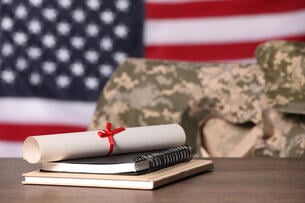 Two notebooks and a rolled-up diploma sit on a table in front of an empty chair draped with an Army jacket. The background of the photo is an American flag.