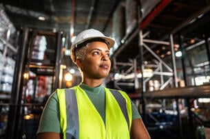 A Black woman in a hard hat and neon vest stands in a warehouse.