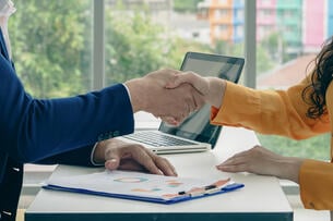 Two people shaking hands in front of a laptop. The laptop is open and has a picture of a person on it.