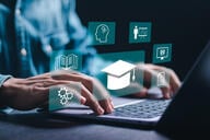 Hands at a computer keyboard with college-type symbols (graduation cap, etc.) popping up in front of the screen (concept image). 