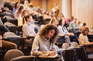 Women sit in a lecture hall. One writes in a notebook. 