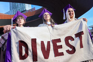 Three CUNY students in purple graduation robes hold a sign that says "Divest."