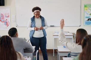 A young college instructor stands in front of a classroom of students; she is listening to a student speak, while another has a hand raised.