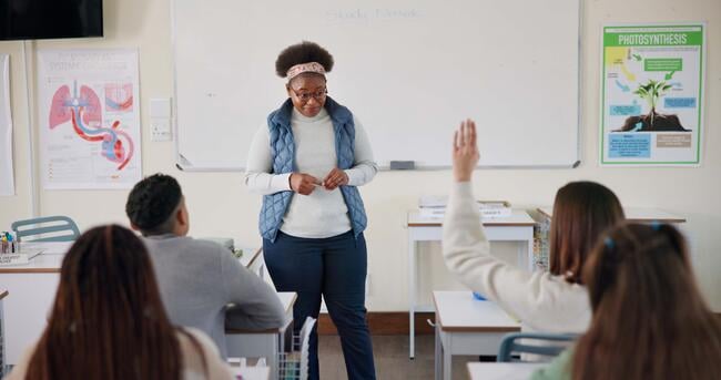 A young college instructor stands in front of a classroom of students; she is listening to a student speak, while another has a hand raised.