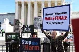 A blonde congresswoman holds up a sign that says there are "two genders, male and female," and "trust the science."