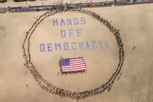 Protesters form a human banner on Ocean Beach in San Francisco; protesters are arranged in a circle around text that says "Hands Off Democracy" over an American flag