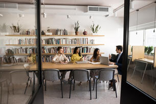 A group of three students can be seen through a doorway talking with a professor at a conference table in a book-lined, light-filled room. 