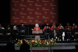 Jane Fonda stands in a red suit in front of a large backdrop displaying USC Annenberg's logo.