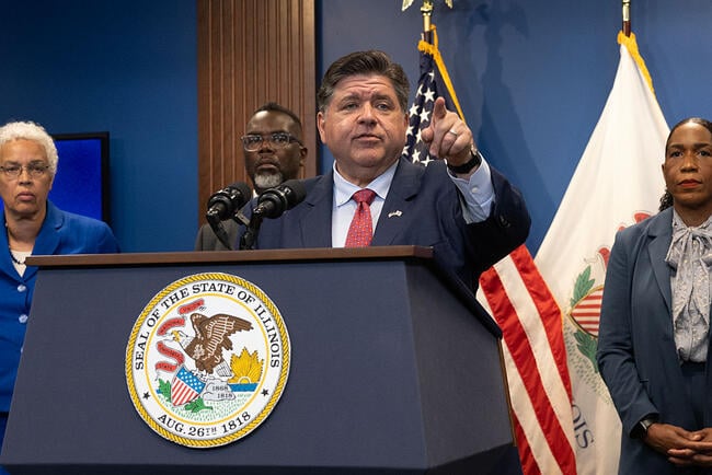 Illinois Governor JB Pritzker stands at a podium, pointing into the distance. He is a white man with dark hair, wearing a blue suit and red tie.