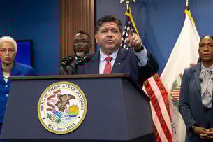 Illinois Governor JB Pritzker stands at a podium, pointing into the distance. He is a white man with dark hair, wearing a blue suit and red tie.
