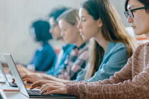 A line of college students of different races and ethnicities works on their laptops in a classroom.