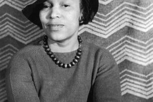 A portrait of author Zora Neale Hurston from around the 1940s. She wears a wide-brimmed hat and smiles while staring directly at the camera.