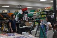 A college student stands behind a video camera, interviewing a subject in the garden section of a store