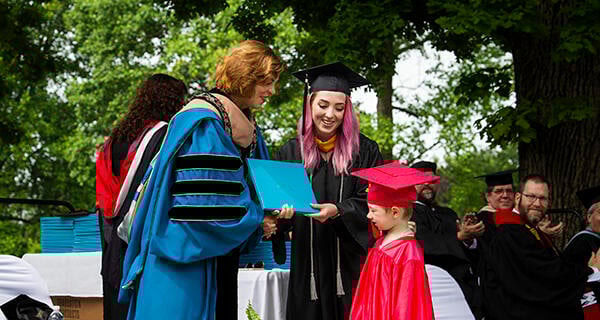 A mother and her young son, both dressed in graduation regalia, accept a diploma on stage from an administrator