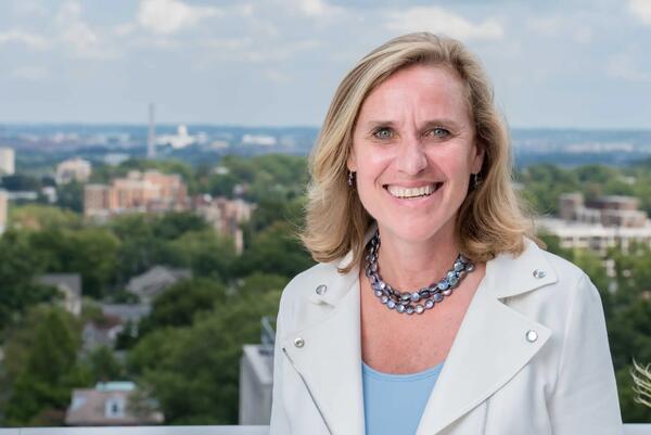 A blonde woman is wearing a white jacket and blue top and standing against the skyline of DC