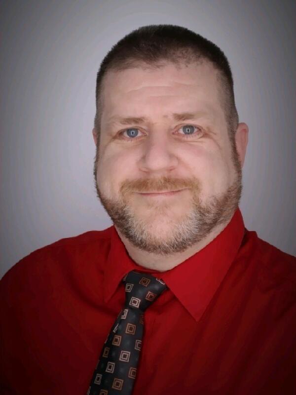 Michael Logan poses for a headshot wearing a red button-up shirt and a black tie. He has short blond hair and facial hair