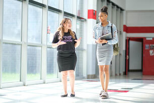 A shorter white woman wearing a shirt that says "college is for everyone" is shown talking with a taller Black girl in a denim jacket and skirt.