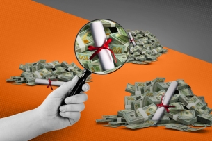 A hand holds a magnifying glass over a diploma sitting on a pile of dollar bills.