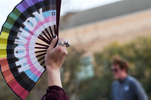 A protester holds up a hand fan in LGBTQ+ pride flag colors with "Howdy" written across it. 