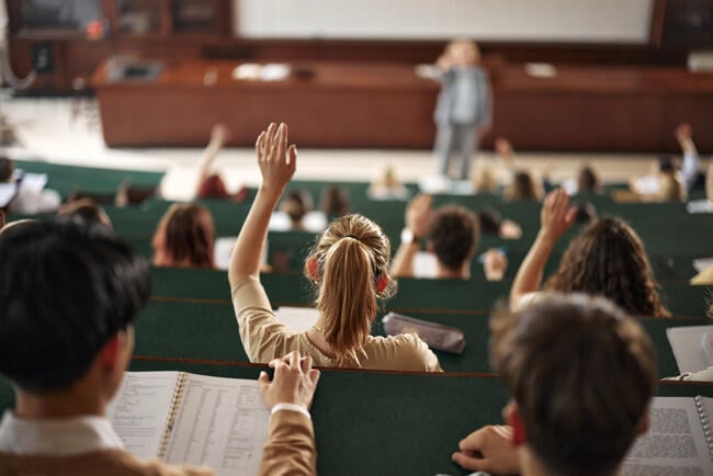 In a lecture hall, a young blonde woman in the middle has her hand raised.