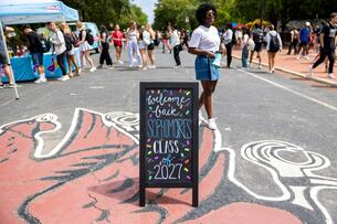 A chalkboard with the text "Welcome back Sophomores, Class of 2027" is in front of an on-campus carnival event 