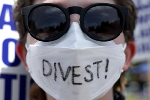 A photograph of the face of a person wearing sunglasses and a white face mask, on which "Divest!" is written. 