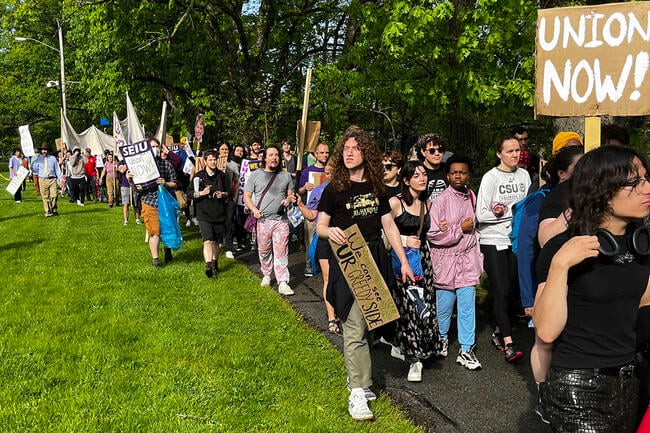 People march outside, some carrying signs, one of which says, "Union Now!"